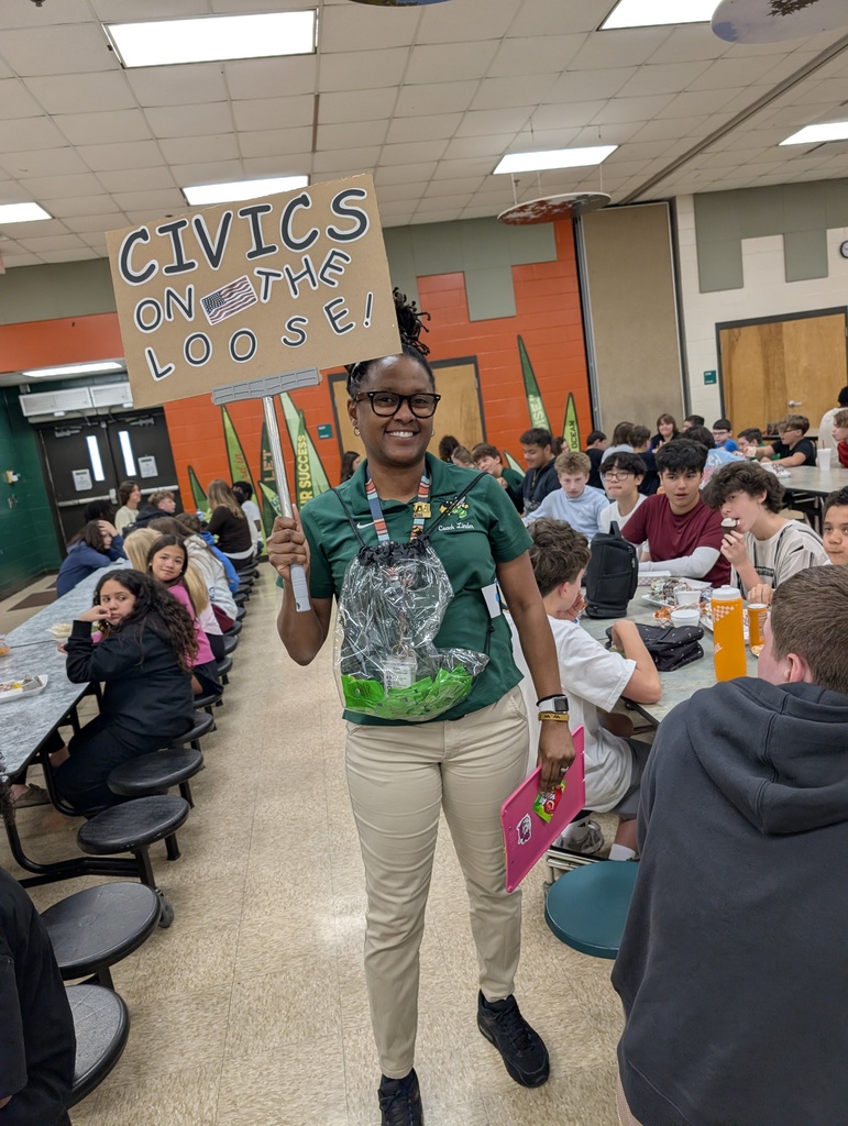 MrsDuncanson, wearing a green shirt and khaki pants, holds a sign that reads, Civics on the Loose! as she walks around the lunch room asking students who are pictured seated at their lunch tables questions about civics and gives out candy to those with correct answers. 