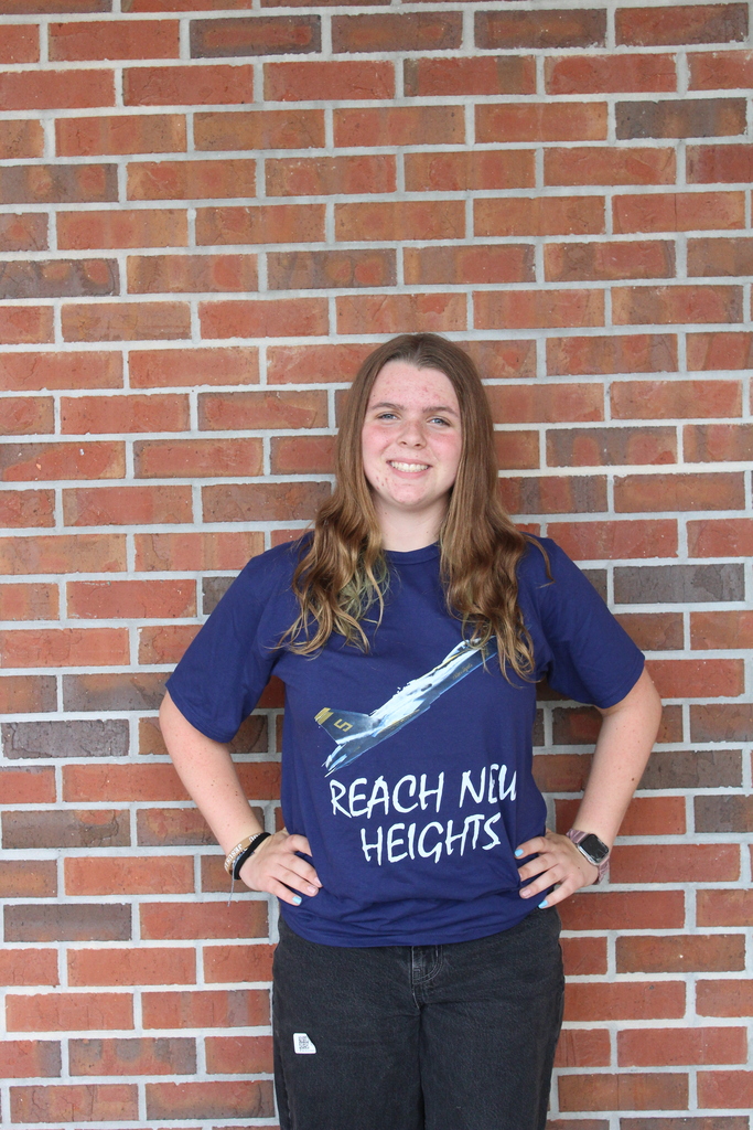 Female student with brown hair poses for the camera in a blue shirt that reads "Reach New Heights" and has an airplane taking off in front of a brickwall with her hands on her hips. 