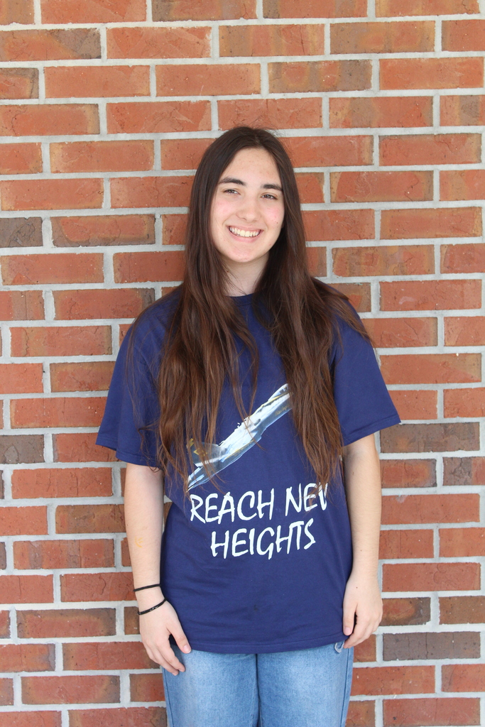 Female student with dark brown hair poses for the camera in a blue shirt that reads "Reach New Heights" and has an airplane taking off in front of a brickwall with her hands by her side.  