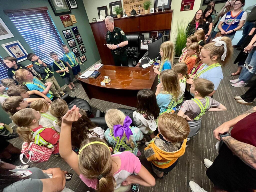Elementary students wearing safety patrol vests stand inside an office at the Nassau County Sheriff’s Office as a law enforcement officer speaks to the group from behind a desk.