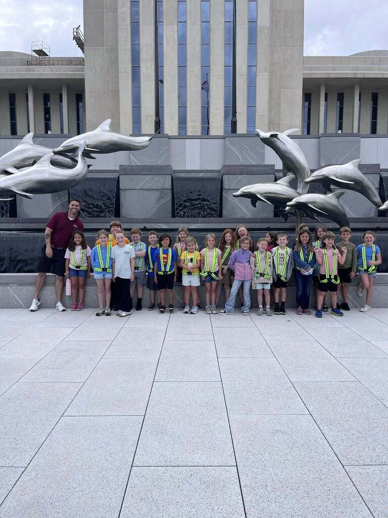 A group of elementary students wearing safety patrol vests stands in front of a dolphin sculpture near the Florida State Capitol.
