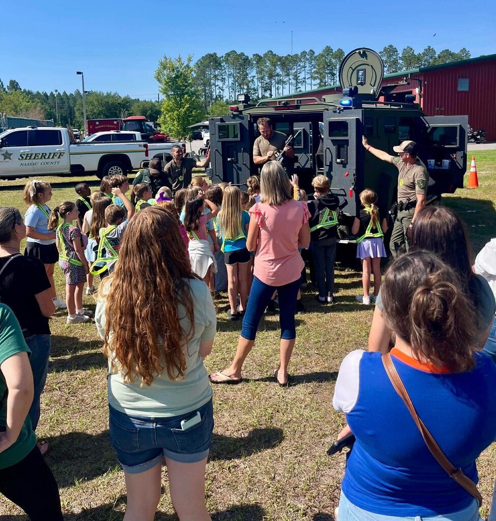 Elementary students and adults gather outdoors as Nassau County Sheriff’s Office personnel demonstrate safety equipment beside an armored vehicle.