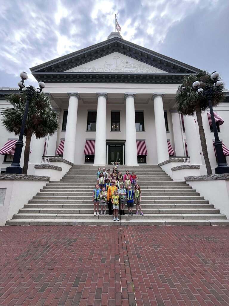 A group of elementary students wearing safety patrol vests stands on the steps in front of the Florida State Capitol building.