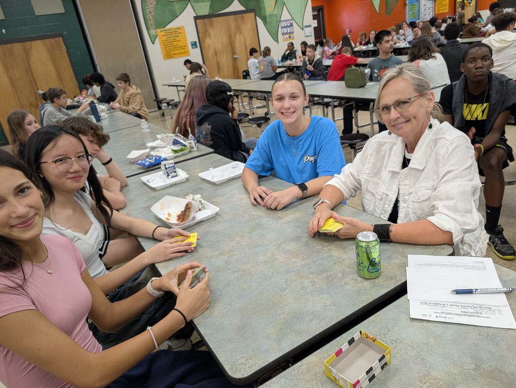 Female teacher smiles for the camera with three of her students as she plays cards with them at the lunch table. 
