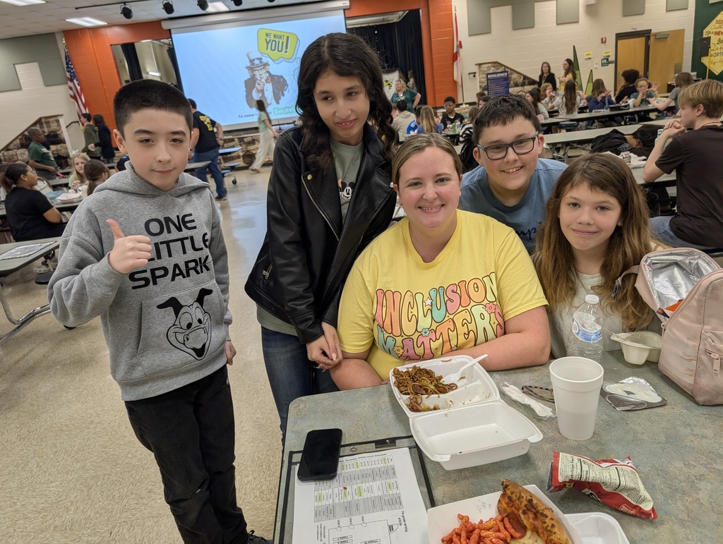 Female teacher smiles for the camera with four of her students at the lunch table. 
