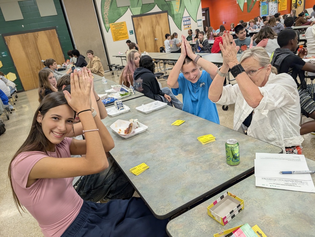 Female teacher acts silly with her students at the lunch table as they put their hands together like a shark fin over their head for the camera. 