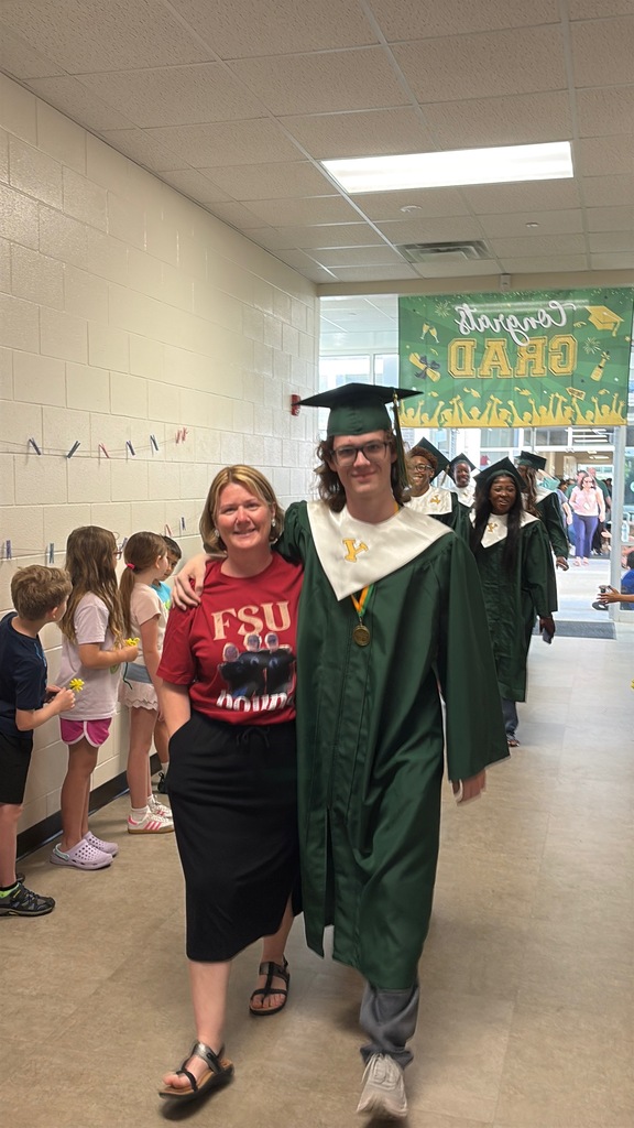 A group of Yulee High School graduating seniors in green caps and gowns walking through a Wildlight Elementary school hallway lined with smiling students cheering them on.