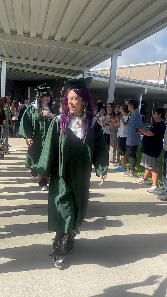A group of high school graduating seniors in green caps and gowns walking through an elementary school hallway lined with smiling elementary students cheering them on.