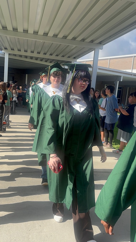A group of high school graduating seniors in green caps and gowns walking through an elementary school hallway lined with smiling elementary students cheering them on.
