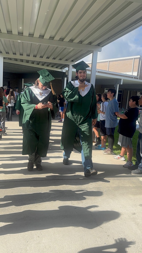 A group of high school graduating seniors in green caps and gowns walking through an elementary school hallway lined with smiling elementary students cheering them on.