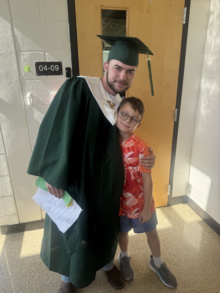 A tall high school graduate in a green cap and gown smiling with his arm around a young elementary school boy wearing a bright orange shirt inside a school hallway.