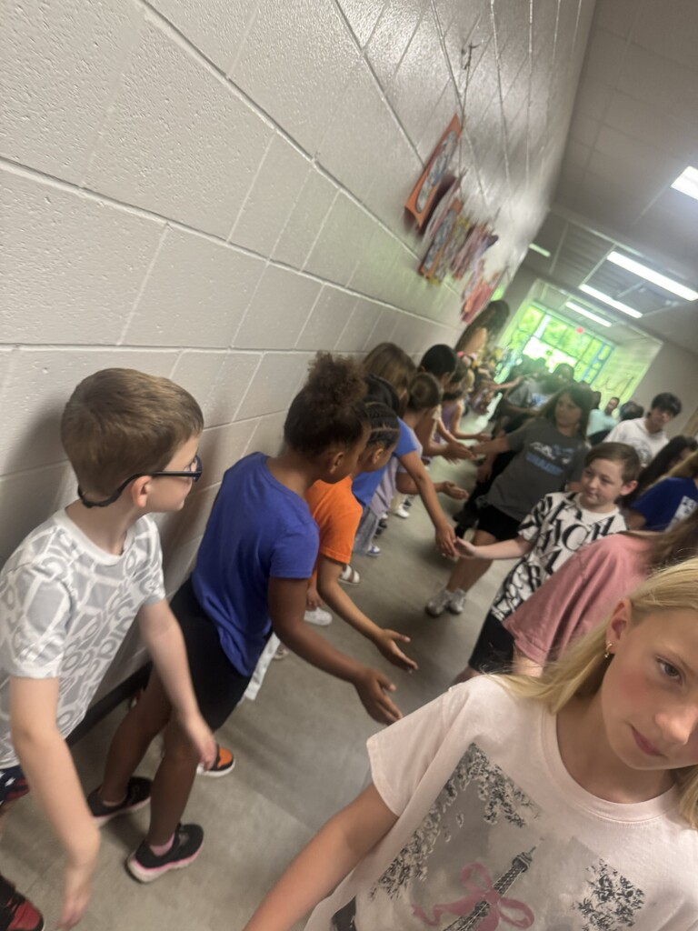 A row of elementary school students standing against a hallway wall, smiling and waiting to cheer for graduating seniors.