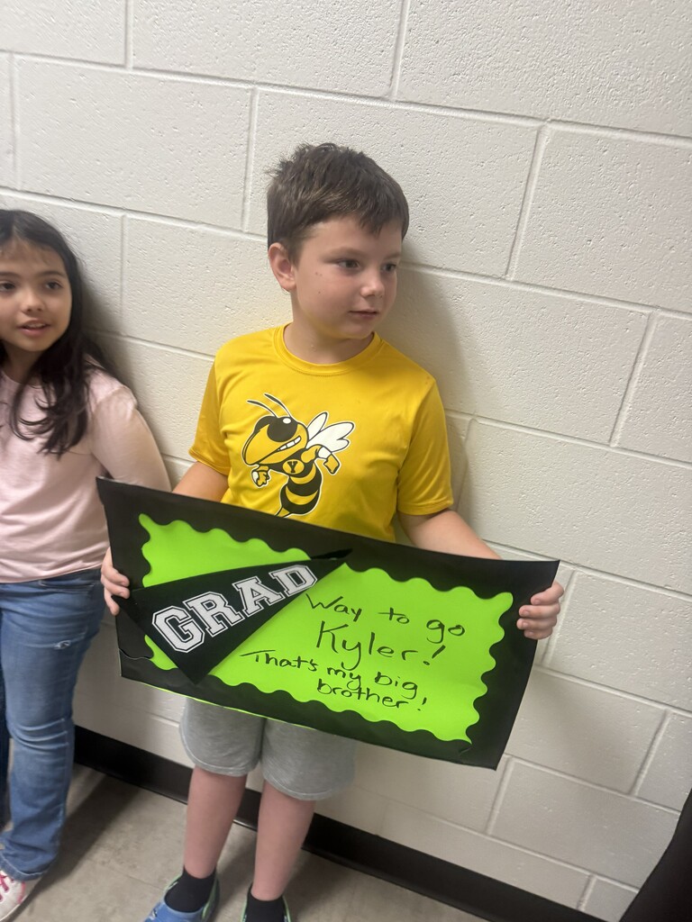 A young elementary school boy in a bright yellow t-shirt and a young girl stand against a school wall, smiling and holding up a green and black handmade graduation sign that reads "Way to go Kyler!"