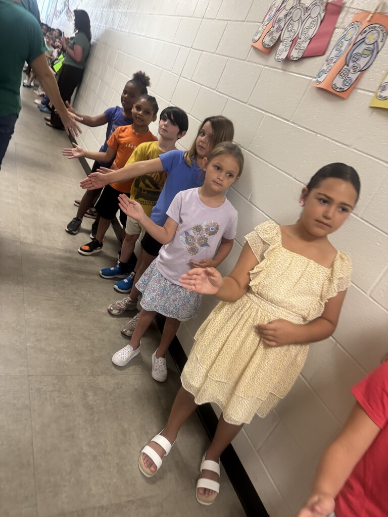 A row of elementary school students standing against a hallway wall, smiling and waiting to cheer for graduating seniors.