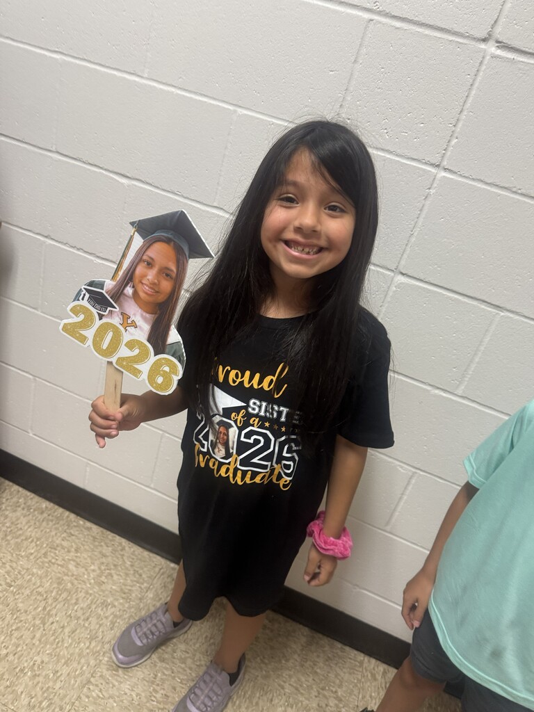 A young elementary school girl in a black shirt stands in a school hallway, smiling and holding a handmade photo prop cutout of a graduate that reads "Class of 2026."