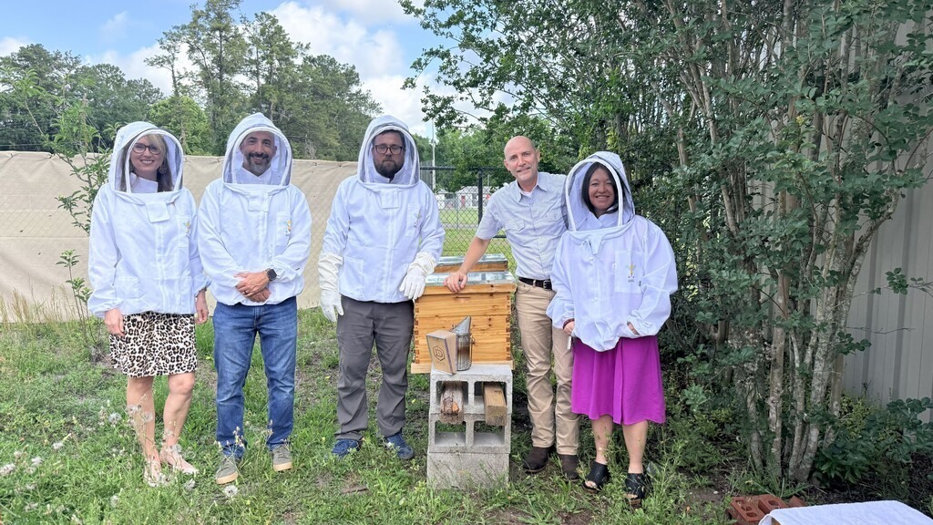 Staff members wearing protective beekeeping suits pose beside beehives during a honey harvesting demonstration at Hilliard Middle-Senior High School.