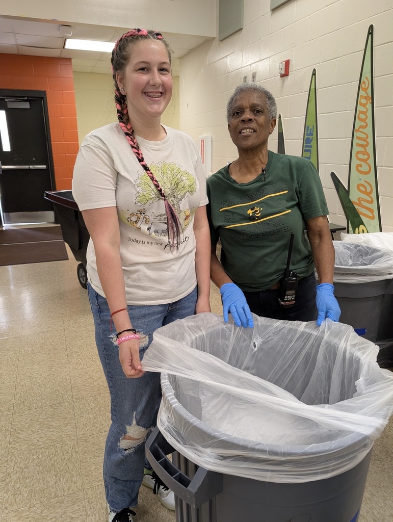 Female custodia with short black and grey hair, wearing blue latex gloves smiles for the camera while holding a trashcan and standing next to a student who is smiling after helping her pick up trash around the cafeteria. 