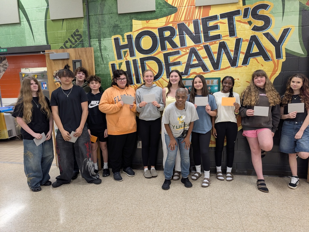 Female custodian leans forward and smiles for the camera with eleven students as they hold up their envelopes that she gave them. Each envelope had a gift card in it thanking them for their behavior in the cafeteria.