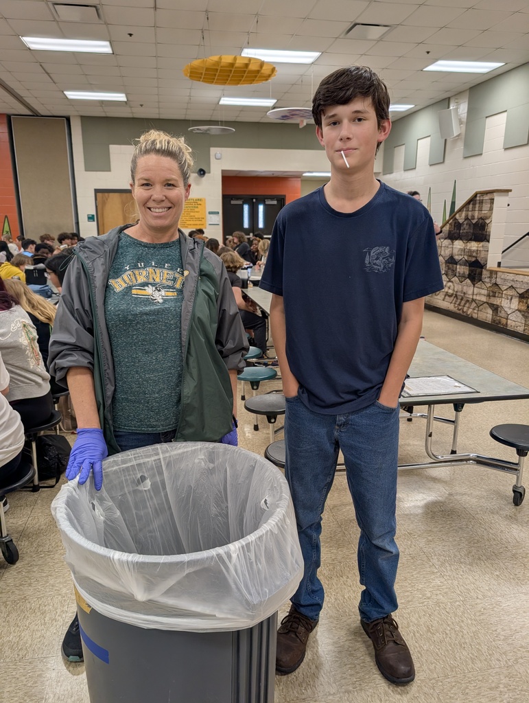 Female custodian with brown hair, wearing blue latex gloves smiles for the camera with a middle school student who has been helping her pick up trash around the cafeteria. 