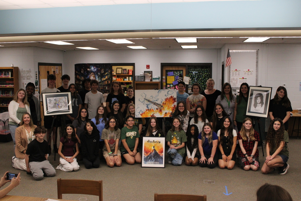Over 20 students kneel and stand while posing for a picture with three of them holding art that they created and a framed photo of the cover of the Resiliency Book in the center. Staff members, including the art teachers, the journalism teacher and the principal of YMS are smiling for the camera in this big group photo taken in the library. 
