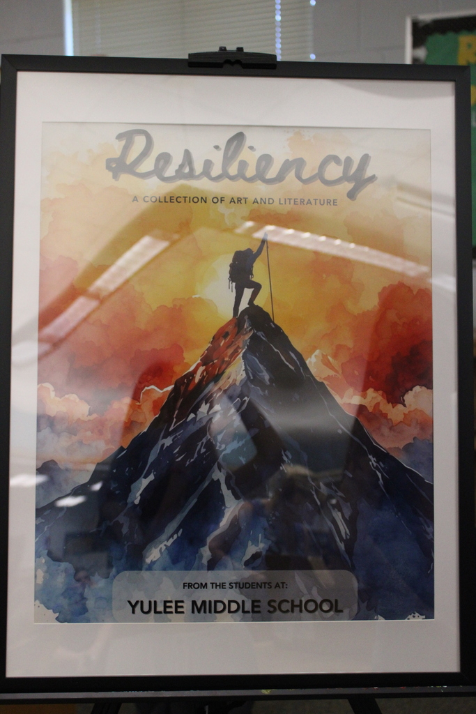 A framed photo of the cover of the Resiliency Book created by the students and staff at Yulee Middle School. The cover is a picture of a mountain climber reaching the summit, behind the climber is yellow, red and white clouds with the sun directly behind the climber.