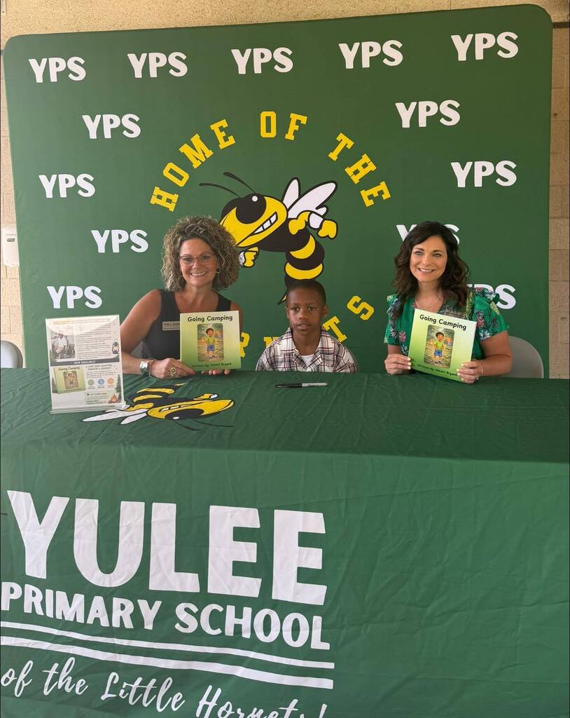 Two Yulee Primary School staff members hold copies of Going Camping while sitting beside first grade student author Amari during a campus book signing celebration.