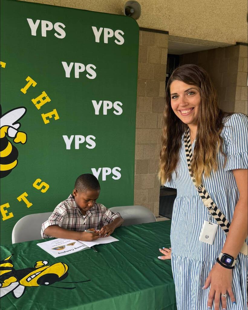 A staff member smiles beside first grade student author Amari as he signs copies of his book Going Camping at a table decorated with Yulee Primary School Hornets branding.