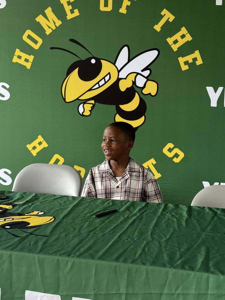 First grade student author Amari smiles while seated at a signing table in front of a green Yulee Primary School Hornets backdrop during his campus book signing event.