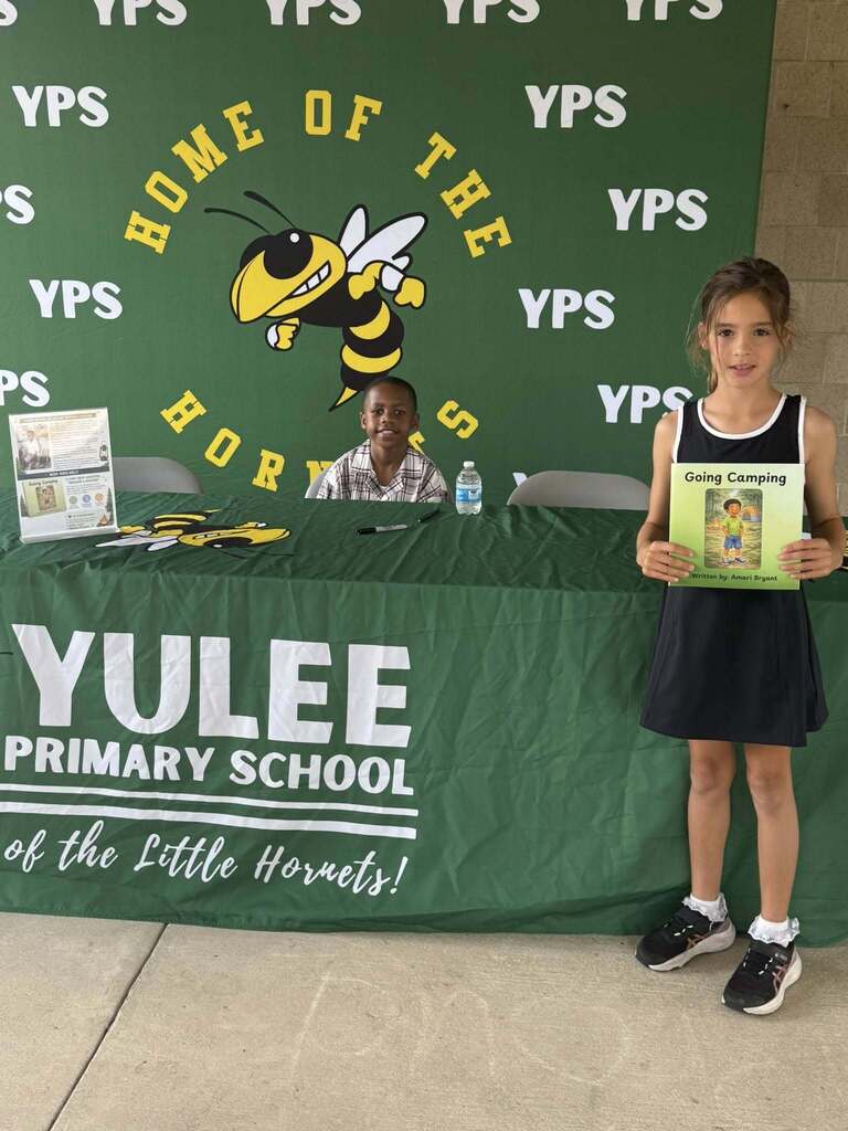 A student stands in front of a Yulee Primary School Hornets backdrop holding a copy of the book Going Camping while first grade student author Amari smiles from behind the signing table during a campus book signing event.