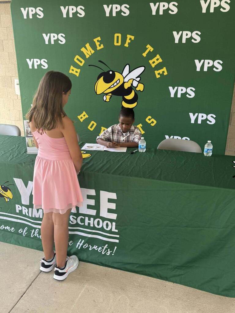A student stands at the signing table while first grade student author Amari signs a copy of his book Going Camping during a Yulee Primary School event.