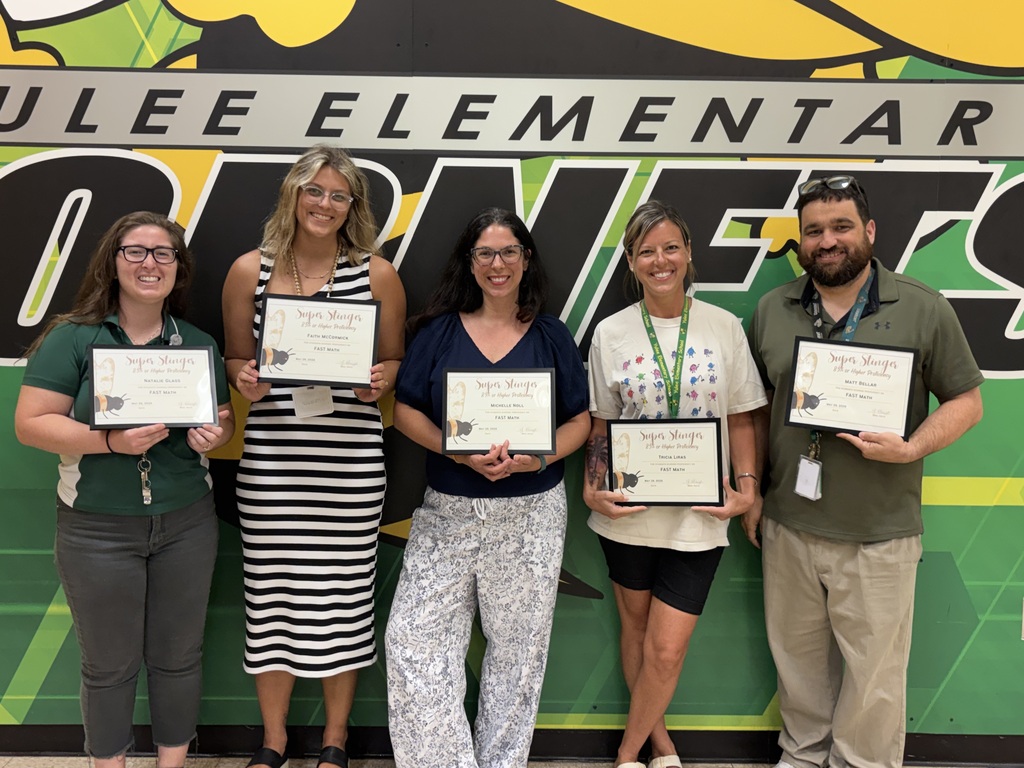 A group photo of Yulee Elementary teachers and staff smiling and holding certificates in front of a green and yellow wall featuring the school's "Hornets" logo and mascot.