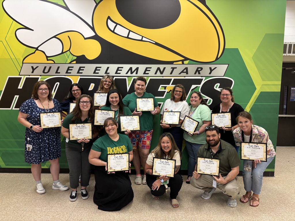 A group photo of Yulee Elementary teachers and staff smiling and holding certificates in front of a green and yellow wall featuring the school's "Hornets" logo and mascot.