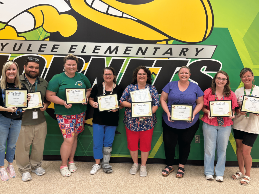 A group photo of Yulee Elementary teachers and staff smiling and holding certificates in front of a green and yellow wall featuring the school's "Hornets" logo and mascot.