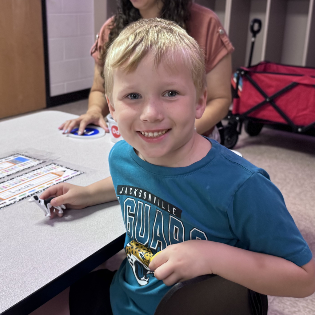 A smiling student sits at a classroom table holding a small toy while classroom materials are arranged in front of them. A staff member is visible in the background.