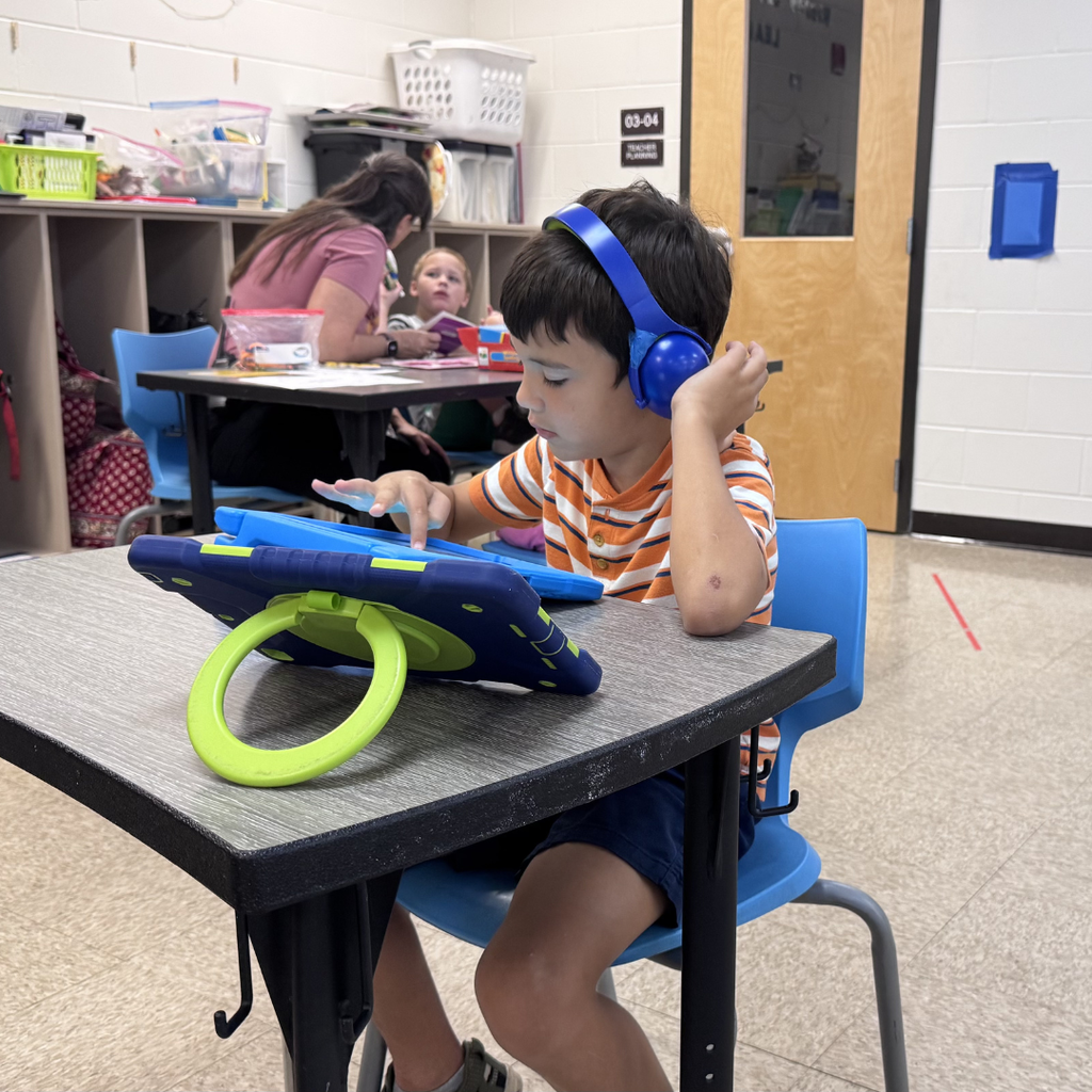 A student wearing blue headphones uses a tablet while seated at a classroom table. Another teacher works with a student in the background, and classroom learning materials are visible throughout the room.