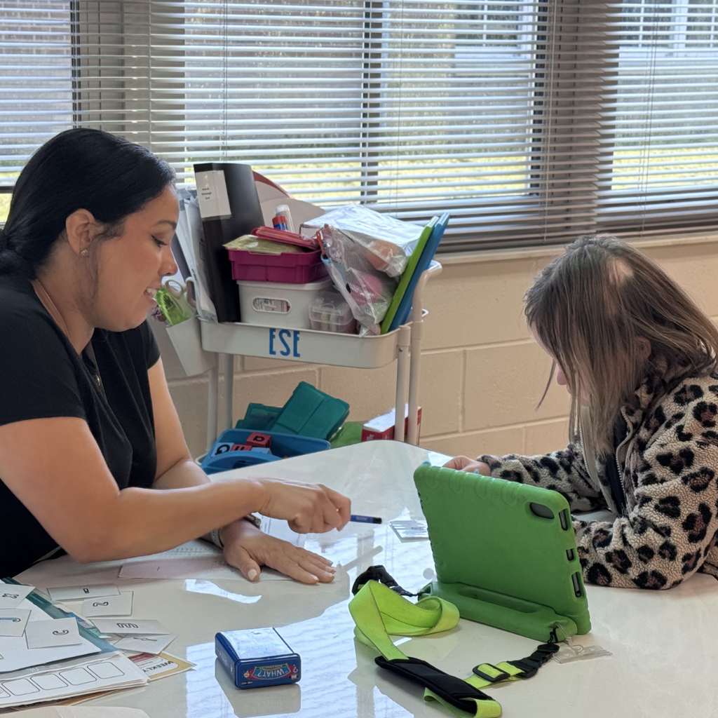 A teacher works one-on-one with a student seated at a classroom table. Learning materials and letter cards are spread across the table as the teacher points to an activity while the student follows along on a tablet.