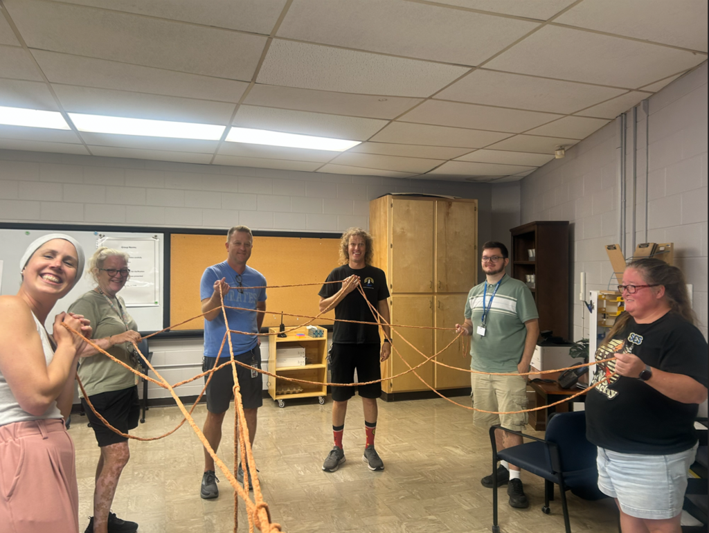 Six Southside Elementary staff members stand in a circle indoors participating in a team-building activity, each holding a section of orange yarn that forms a connected web between them. The group is smiling and engaged, representing collaboration, communication, and the importance of every team member's role in building strong connections. The activity takes place in a classroom or meeting space with cabinets, a bulletin board, and office furniture visible in the background.