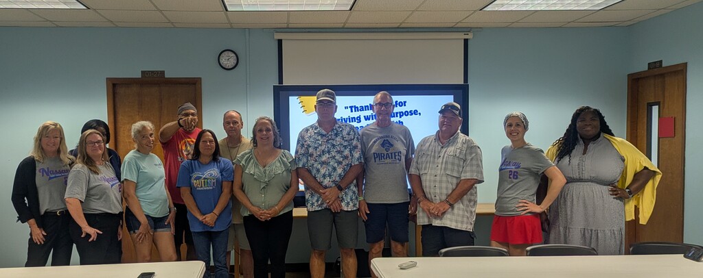A group of approximately 12 school bus drivers and school administrators stand together smiling in a meeting room for a group photo. A presentation screen behind them displays a welcome message thanking bus drivers for "driving with purpose." The group is dressed in a mix of school spirit shirts and casual attire, reflecting a welcoming, team-oriented atmosphere as they gather to prepare for the new school year. Tables are visible in the foreground, and the room includes classroom-style features such as a projector screen, clock, and doors.