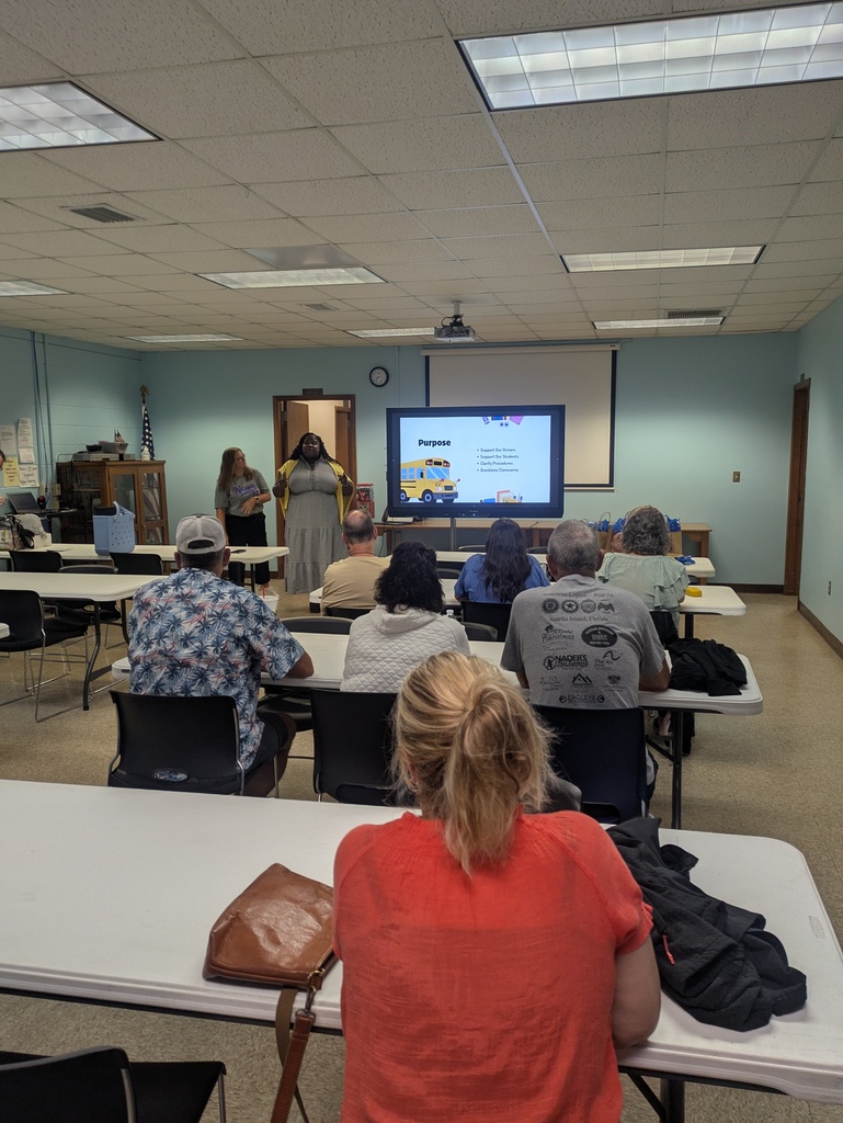School administrators stand at the front of a meeting room presenting to a group of school bus drivers seated at tables during a welcome-back meeting. A large presentation screen displays a slide titled "Purpose" with a school bus graphic and key discussion points about supporting bus drivers, supporting students, clarifying procedures, and addressing questions or concerns. The attendees are focused on the presentation, creating a collaborative and professional atmosphere as the team prepares for the upcoming school year. The room includes classroom-style tables and chairs, an American flag, and presentation equipment.