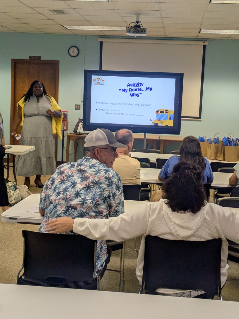 A school administrator stands at the front of a meeting room leading an interactive discussion with a group of school bus drivers during a welcome-back meeting. A large presentation screen displays the activity titled "My Route...My Why," encouraging drivers to reflect on what they enjoy most about being a bus driver and when they know they have made a difference. Bus drivers are seated at tables, engaged in conversation and smiling as they participate. Gift bags are lined up on a table near the front of the room, adding to the welcoming atmosphere as the transportation team prepares for the new school year.