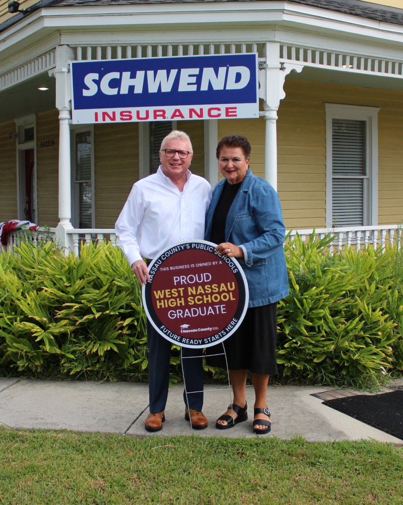 Paul Schwend, West Nassau High School graduates and owners of Schwend Insurance Agency Inc., stand outside their insurance office holding a Nassau County School District Alumni Business Recognition sign that reads, "Proud West Nassau High School Graduate." The recognition celebrates alumni-owned businesses and the lasting impact of Nassau County's public schools.
