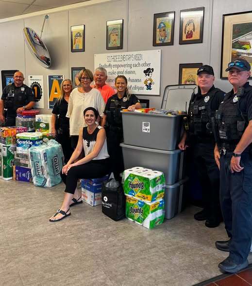 A group of community members, school staff, and local law enforcement officers stand together inside Southside Elementary School with donated supplies, including large storage bins, paper towels, toilet paper, cleaning supplies, snacks, and other essential items. A seated woman smiles in front of the donations while others stand behind them. Student artwork and the Southside Elementary mission statement are displayed on the wall in the background, highlighting the school's welcoming environment and strong community partnerships.