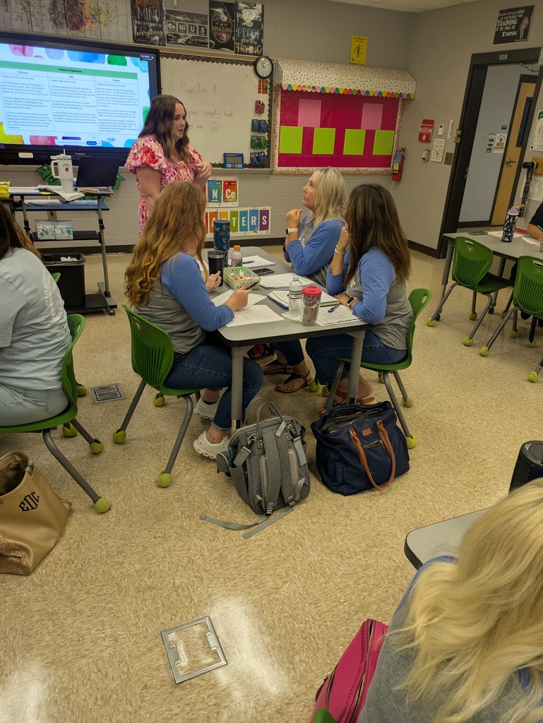 Teachers participate in a professional learning session as an instructor leads a discussion at the front of the classroom while small groups collaborate at their tables.