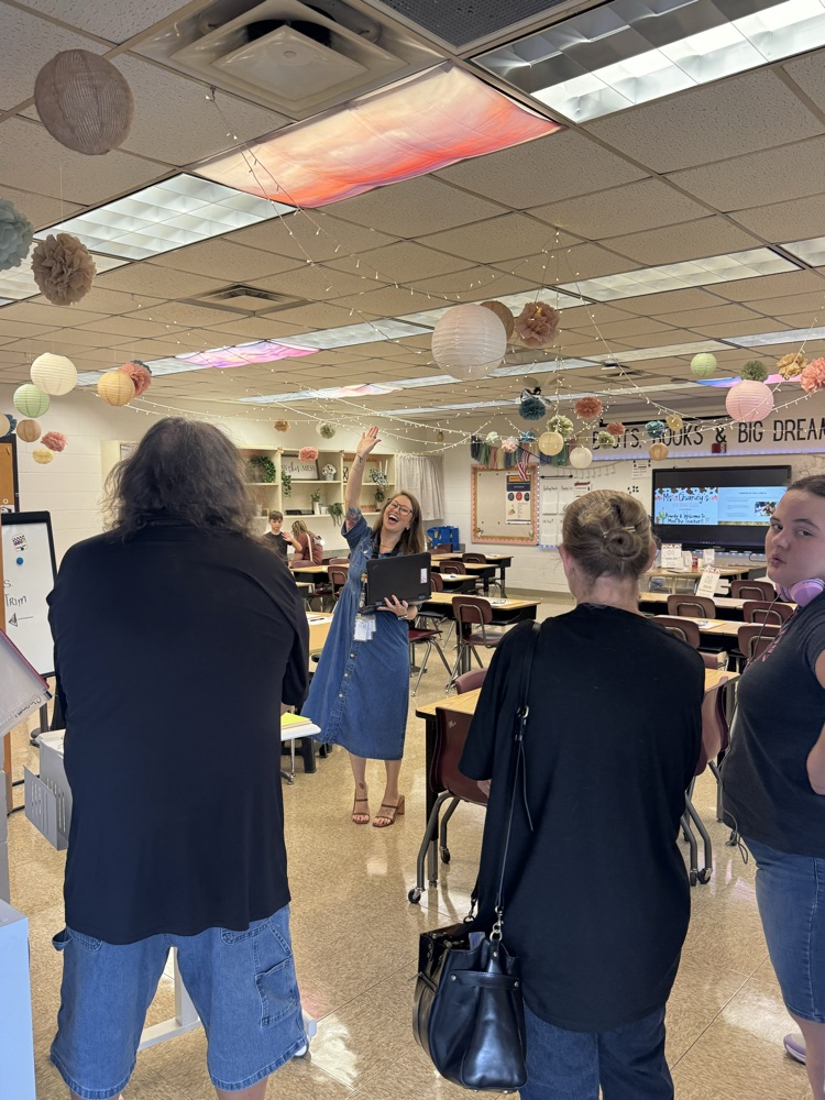 Ms. Chaney welcomes students and their families into a decorated classroom during a back-to-school meet-and-greet. The teacher smiles, waves, and holds a laptop while speaking with visitors. 
