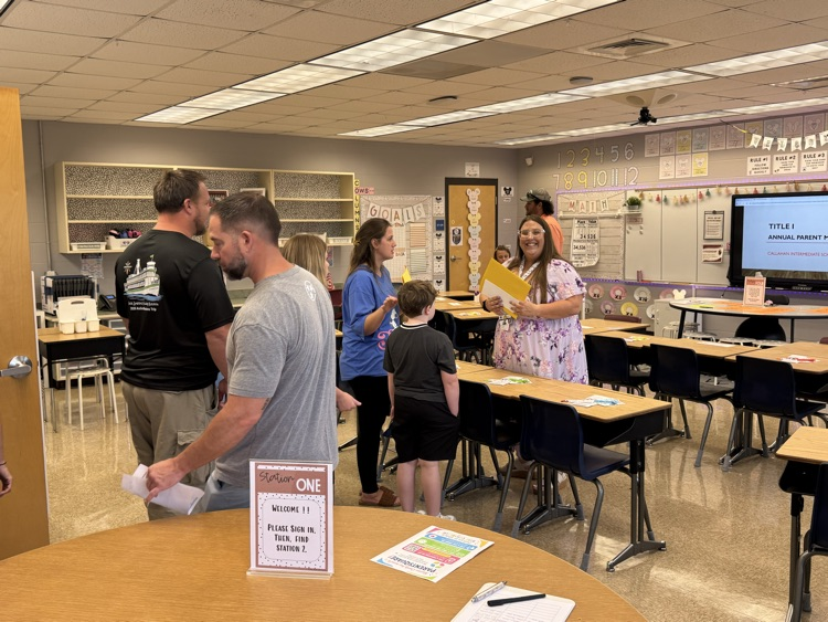 Ms. Williams greets students and their families in her classroom during a back-to-school meet-and-greet while holding student folders and answering questions.