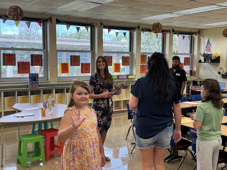 Mrs. Aspinwall welcomes students and their families during a back-to-school meet-and-greet in her classroom while holding student materials. A students waves towards the camera.