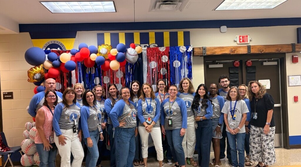 A group of Southside Elementary educators poses together in front of a festive baseball-themed backdrop decorated with blue, red, and gold balloons, streamers, and baseballs. The staff members wear Southside shirts and smile for the group phot