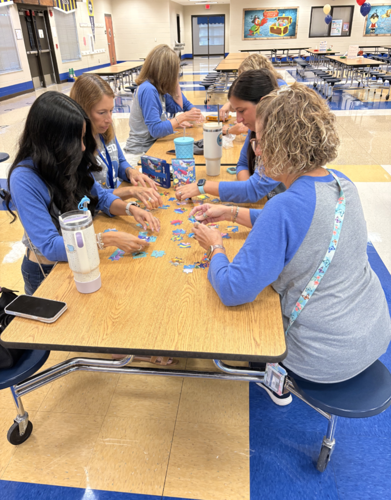 A group of educators sits around a cafeteria table working together on colorful jigsaw puzzles during a team-building activity. The school’s blue-and-gold colors and pirate-themed decorations are visible in the background.