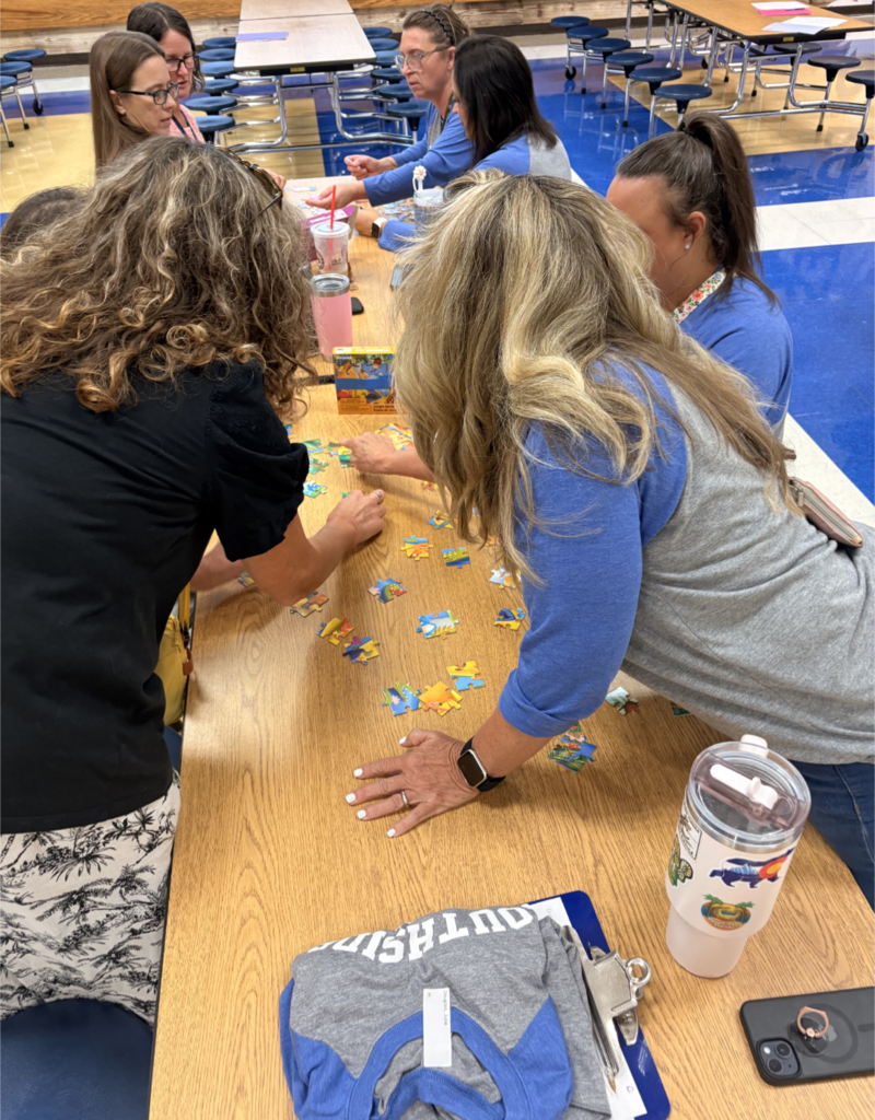 Several educators gather around a cafeteria table working together to assemble colorful jigsaw puzzles as part of a collaborative team-building activity.