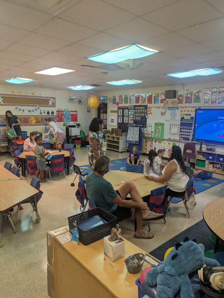 A classroom filled with students, families, and educators gathered around tables for an activity. Colorful learning displays, a large digital screen, classroom materials, and a blue-and-yellow rug are visible throughout the room.